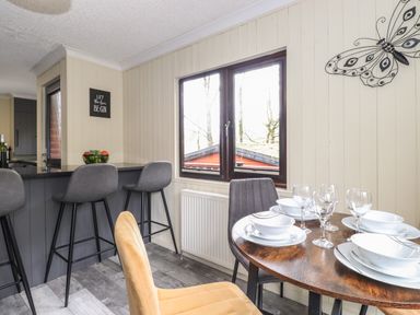 A dining area with a round wooden table set with plates and glasses next to a window and a breakfast bar with three grey stools at Alfresco Lodge Skiptory Howe 37B White Cross Bay near Windermere