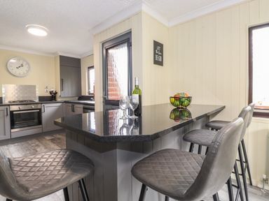 A kitchen with a black countertop bar with three chairs a fruit bowl and wine bottle at Alfresco Lodge in Skiptory Howe 37B White Cross Bay near Windermere