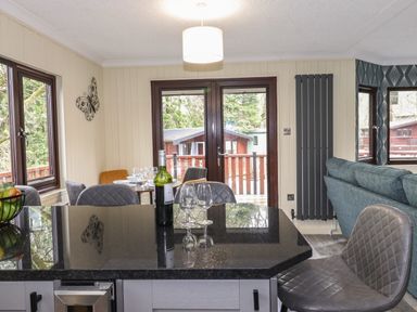 A dining and living area with a black countertop island a bottle of wine and wine glasses a blue sofa and double doors leading outside at Alfresco Lodge Skiptory Howe 37B White Cross Bay near Windermere