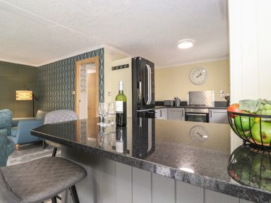 A kitchen with a black countertop, chairs, a wine bottle, glasses, a fruit bowl, and a view of a living area at Alfresco Lodge in Skiptory Howe 37B White Cross Bay near Windermere