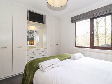 A bedroom with a double bed and green throw next to a window with gray blinds and built in wardrobes with a mirror and a tv at Alfresco Lodge Skiptory Howe 37B White Cross Bay near Windermere