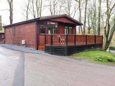 A wooden cabin with a raised deck and railing surrounded by trees at Alfresco Lodge Skiptory Howe 37B White Cross Bay near Windermere