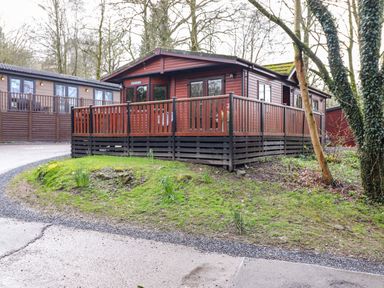 A wooden cabin with a fenced porch on a grassy area near a paved path at Alfresco Lodge in Skiptory Howe 37B White Cross Bay near Windermere