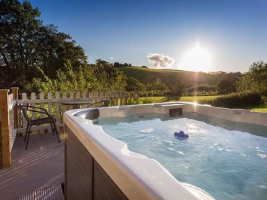 An outdoor hot tub on a deck with two chairs and a table overlooking a grassy hill at Apple Barn in South Molton