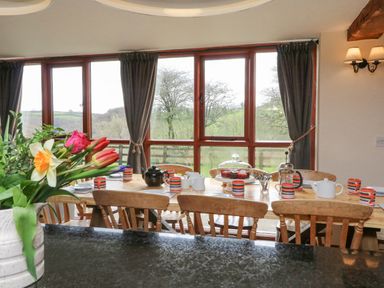 A dining room with a wooden table set with teapots mugs and a cake stand near large windows with curtains at Apple Barn in South Molton