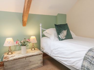 A bedroom with a single bed with white bedding and a green pillow next to a wooden chest with two lamps and a vase with flowers at Apple Barn in South Molton