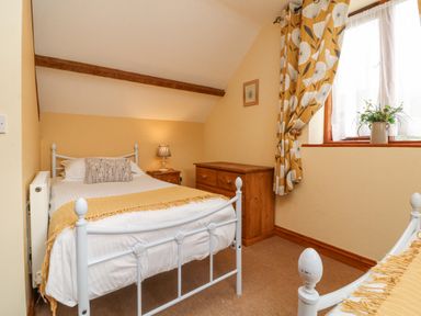 A small bedroom with a white metal bed a wooden dresser a wooden nightstand with a lamp and a window with patterned curtains at Apple Barn in South Molton