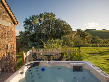 An outdoor hot tub next to a stone building with a small table and two chairs behind a white picket fence at Apple Barn in South Molton