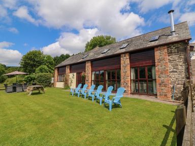 A stone and brick building with large windows and a row of blue chairs on the lawn with a picnic table and outdoor dining set at Apple Barn in South Molton