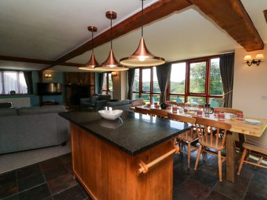 A dining area with a wooden table set with cups and a kitchen island with hanging lights and sofas in the background at Apple Barn in South Molton