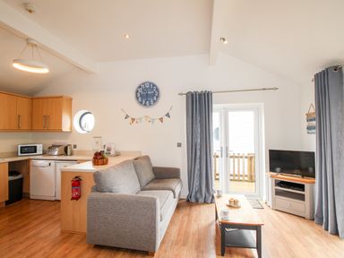 A living room and kitchen area with a grey sofa wooden cabinets a clock and a TV at Beside the Sea in Eype