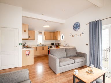 An open plan kitchen and living area with grey sofas wooden cabinets and a wooden coffee table with mugs and pastries at Beside the Sea in Eype