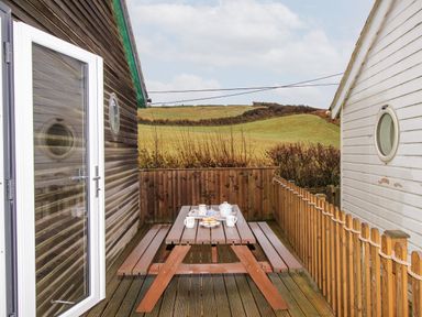 An outdoor wooden picnic table set with cups and a teapot between two buildings with views of grassy hills at Beside the Sea in Eype
