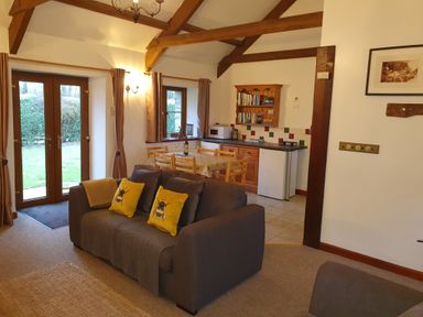 A living area with a gray sofa with yellow pillows a dining table with chairs a small kitchen with wooden cabinets a bookshelf a microwave and double glass doors at Ty Barcud in Llanfair Clydogau