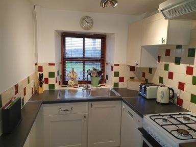 A kitchen with a sink under a window a clock on the wall tiled backsplash a kettle toaster stove and utensils at Ty Barcud in Llanfair Clydogau