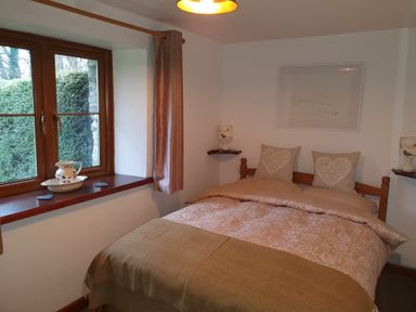 A bedroom with a double bed two heart patterned pillows two small shelves with lamps and a window with curtains at Ty Barcud in Llanfair Clydogau
