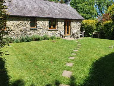 A stone cottage with wooden windows and doors a patio with table and chairs a stone path on the grass and bushes surrounding the lawn at Ty Barcud in Llanfair Clydogau