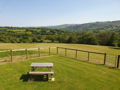 A picnic table on grass in a fenced yard with rolling hills and trees in the background at Ty Barcud in Llanfair Clydogau