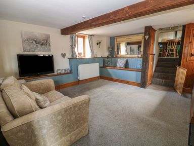 A living room with a patterned sofa a wall mounted television cushions on a built in bench a window and stairs leading to a dining area at Hazel Barn North Molton