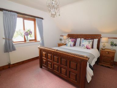 A bedroom with a wooden bed frame floral pillows two bedside tables with lamps and flowers a window with blue curtains at Hazel Barn in North Molton
