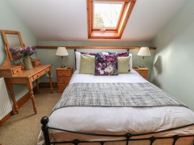 A bedroom with a bed under a skylight window two bedside tables with lamps and a wooden dressing table with flowers at Hazel Barn in North Molton