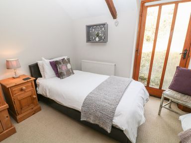 A bedroom with a single bed with white bedding and cushions a wooden bedside table with a lamp a wooden chair with a cushion and a wooden door in Hazel Barn in North Molton