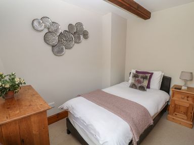 A bedroom with a single bed decorative pillows a wooden nightstand with a lamp a wooden dresser with a plant and metal wall art at Hazel Barn North Molton