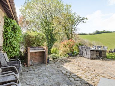 An outdoor patio with wicker chairs a brick barbecue and a dining table set with dishes surrounded by trees and greenery at Hazel Barn in North Molton