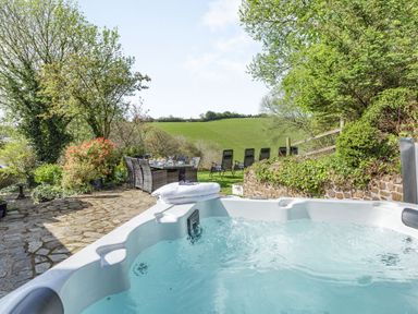 An outdoor area with a hot tub a dining table with chairs and lounge chairs on grass with trees and hills at Hazel Barn in North Molton