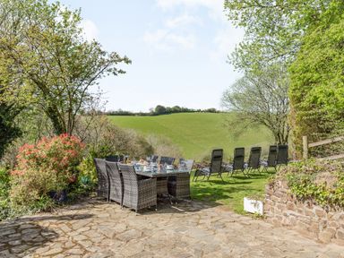 An outdoor patio with a dining table and chairs set with dishes and several lounge chairs on grass overlooking green fields at Hazel Barn in North Molton