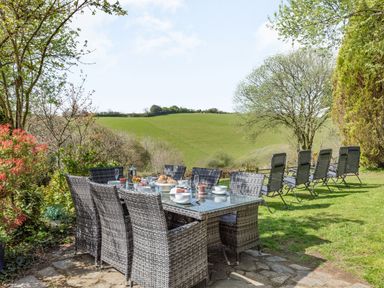 An outdoor dining table with wicker chairs and tableware on a lawn with reclining chairs and trees at Hazel Barn in North Molton