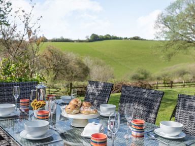 An outdoor dining table set with bowls mugs a teapot and pastries with chairs and a green field in the background at Hazel Barn in North Molton
