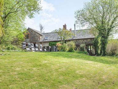 A stone house with outdoor chairs and table on a grassy lawn surrounded by trees at Hazel Barn in North Molton