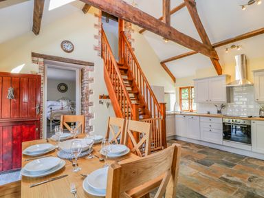 A kitchen and dining area with wooden beams a stairway and a bedroom visible through a doorway at Oak Cottage in North Molton