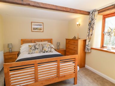 A bedroom with a wooden bed frame two bedside tables with lamps a chest of drawers and a window with patterned curtains at Oak Cottage in North Molton