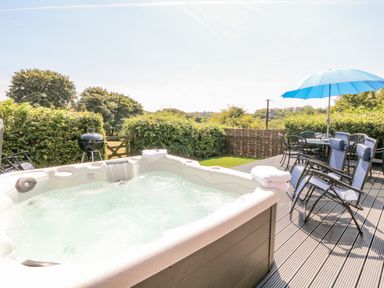 An outdoor deck with a hot tub, two reclining chairs with towels, a dining table with chairs, and a blue umbrella at Oak Cottage in North Molton