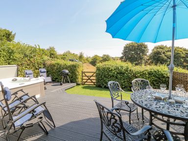 An outdoor patio with metal table and chairs under a blue umbrella a hot tub reclining chairs a grill and a wooden gate at Oak Cottage in North Molton