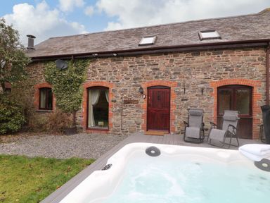 An outdoor area with a white jacuzzi a wooden deck with two gray reclining chairs and a stone building with red brick window frames at The Hawthorns in North Molton