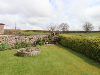 A garden with a stone planter on grass and a wooden bench near a stone wall and hedge at Crablake Farmhouse in Exminster