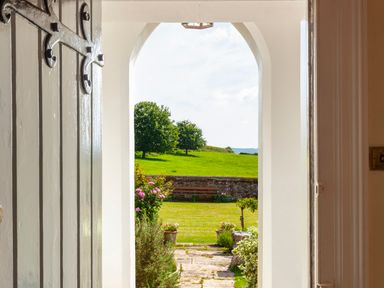 A view through an open door to a garden with flowers a stone path a bench and green fields at Crablake Farmhouse in Exminster