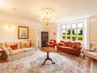 A living room with patterned sofas an armchair a round wooden table a bookshelf and a large window at Crablake Farmhouse in Exminster