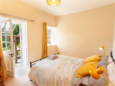A bedroom with a bed yellow pillows and lamps an open door and a window with yellow curtains at Crablake Farmhouse in Exminster