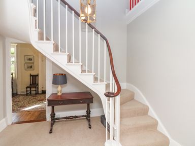A staircase with beige carpet and white railing with a dark wooden handrail a wooden table with a lamp underneath the stairs in a hallway at Crablake Farmhouse in Exminster