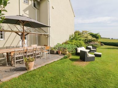 An outdoor patio area with a wooden dining table, chairs, and an umbrella next to a lawn with cushioned wicker chairs and potted plants at Crablake Farmhouse in Exminster
