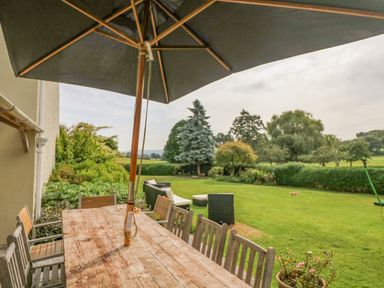A wooden outdoor dining table with chairs and a large umbrella next to a lawn with garden furniture and trees at Crablake Farmhouse in Exminster