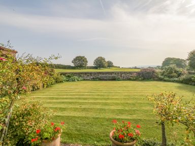 A garden with a striped lawn surrounded by plants and flowers with trees and a bench in the background at Crablake Farmhouse in Exminster