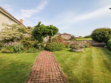 A garden with a brick pathway leading to an archway covered in greenery surrounded by plants and a lawn at Crablake Farmhouse in Exminster
