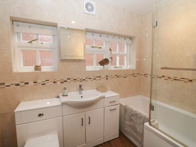 A bathroom with a sink, bathtub with glass screen, toilet, tiled walls, and two windows at The Nook in Whitby