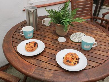 A wooden round table with two mugs, two plates with pastries, a metal coffee pot, and a potted fern at The Nook in Whitby