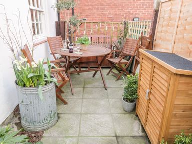 An outdoor patio with a round wooden table and wooden chairs a metal planter with white flowers a wooden storage unit and potted plants at The Nook in Whitby